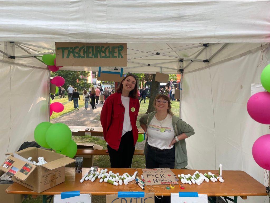 Katharina Schmecht und Alyssia Schröder stehen hinter einem braunen Tisch, auf dem weiße Taschenaschenbecher (Röhrchen) liegen. Der Stand ist ein einem weißen Pavillon